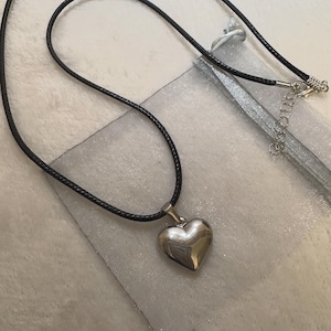 Puede incluir: Un collar con colgante de corazón plateado y cordón negro. El corazón está pulido y es reflectante. El collar se muestra en una bolsa plateada.