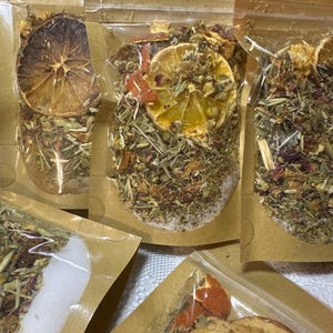 May include: Several small, clear plastic bags filled with dried herbs, flowers, and citrus slices. The bags are sealed and set against a brown paper background. The contents include dried orange and lemon slices, and various green and red herbs.