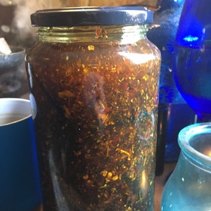 May include: A glass jar filled with a dark brown liquid and various herbs and spices. The jar has a black lid and is sitting on a wooden surface.