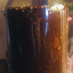 May include: A clear glass jar filled with a dark brown liquid, possibly a sauce or condiment. The liquid contains visible flakes of red pepper and other spices. The jar has a black lid and is likely sealed for preservation.