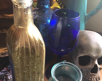 May include: A clear glass bottle with a gold glitter interior and a silver pour spout, a blue sequined bottle, a dark blue wine glass, a light blue glass lantern with a metal handle, and a realistic human skull replica made of grey material.