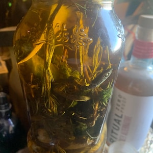 May include: A clear glass jar filled with a golden liquid and various green herbs. The jar has a green lid with the word "Organic" printed on it. The jar has the number "1835" etched on the side.