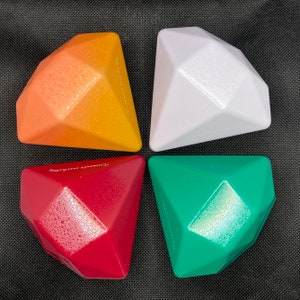 Op de afbeelding: Vier diamantvormige speelgoed in oranje, wit, rood en groen. Het speelgoed is gemaakt van getextureerd plastic.