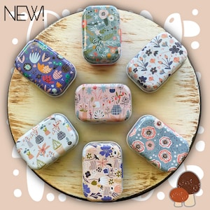 May include: Six rectangular metal tins with rounded corners, each featuring a unique floral or botanical pattern in various colors including pink, blue, purple, and green. The word NEW! is printed in the top left corner of the image.