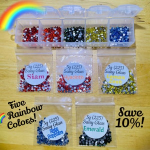 Puede incluir: Un organizador de plástico transparente lleno de pedrería de colores rojo, naranja, negro, amarillo y azul. Cinco bolsas de pedrería están etiquetadas como "Siam", "Hyacinth", "Lemon Zest", "Light Sapphire" y "Emerald". El texto dice "Five Rainbow Colors!" y "Save 10%!"