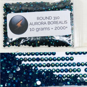 May include: A close-up of a bag of small, round, dark blue iridescent rhinestones. The bag has a label that reads "ROUND 310 AURORA BOREALIS 10 grams = 2000+".