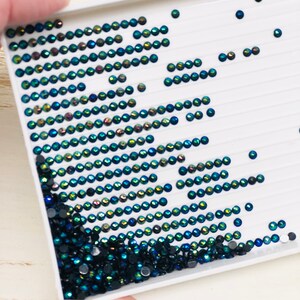 May include: A white tray with rows of small, iridescent green and black rhinestones. The rhinestones are arranged in a diagonal pattern, with the green rhinestones forming a wave-like shape.