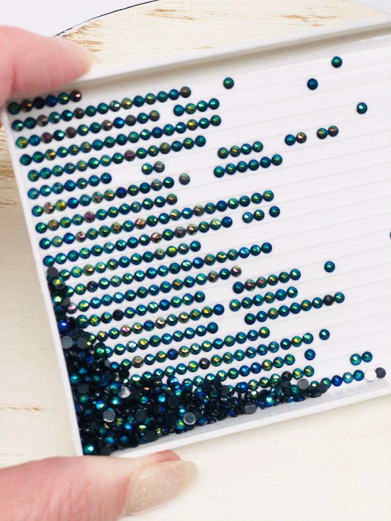 May include: A white tray with rows of small, iridescent green and black rhinestones. The rhinestones are arranged in a diagonal pattern, with the green rhinestones forming a wave-like shape.