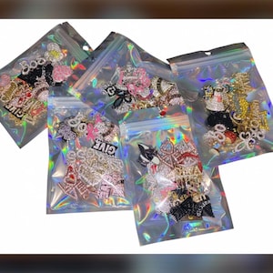 May include: Five clear holographic resealable plastic bags filled with an assortment of decorative metal charms. The charms feature various shapes, including letters, crowns, and symbols, many embellished with small sparkling rhinestones.