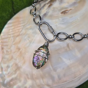 May include: A necklace featuring a silver-toned metal chain with oval links and a screw-lock carabiner clasp. An oval-shaped, iridescent abalone shell pendant is wrapped in thin silver-toned wire and attached to the carabiner clasp.