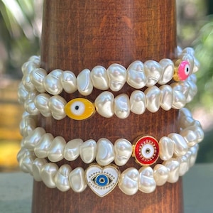May include: Four pearl bracelets with gold accents and evil eye charms. The charms are in various colors, including yellow, pink, red, and blue. The bracelets are stacked on a wooden stand.