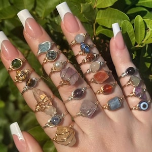 May include: A selection of gemstone rings, each with a unique stone and wire wrapping. The rings include amethyst, quartz, and pearls, set in gold and silver wire. The background is green foliage.
