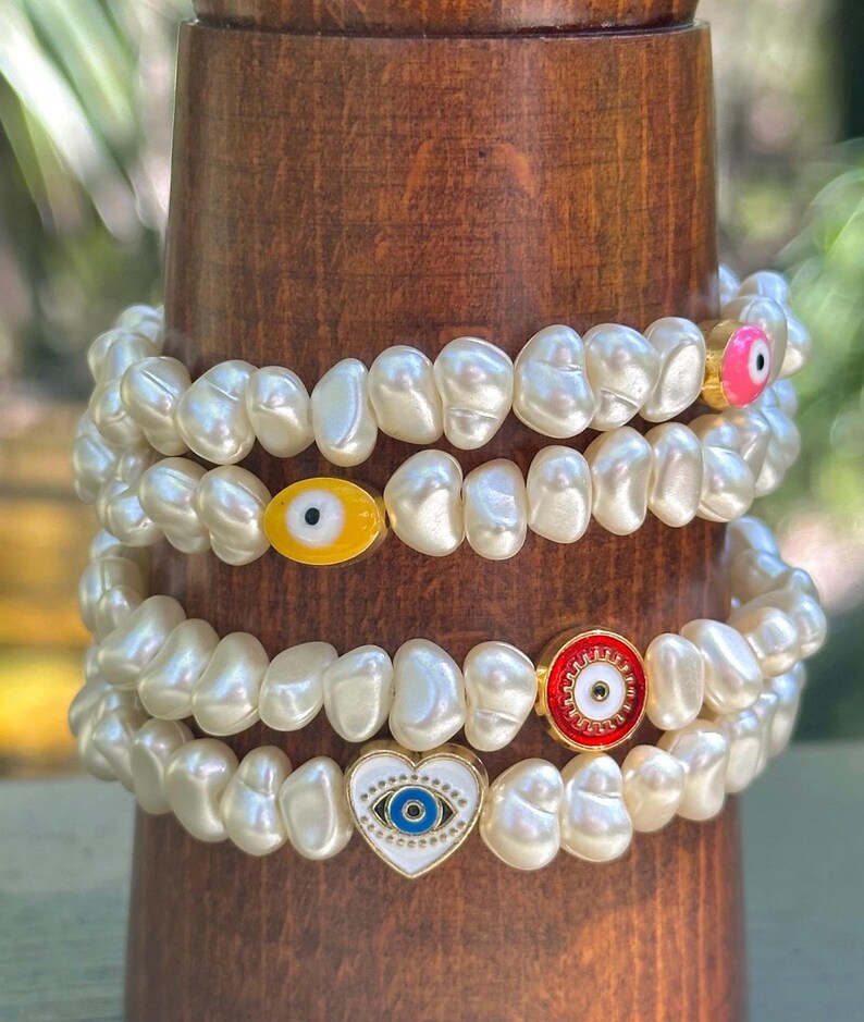May include: Four pearl bracelets with gold accents and evil eye charms. The charms are in various colors, including yellow, pink, red, and blue. The bracelets are stacked on a wooden stand.