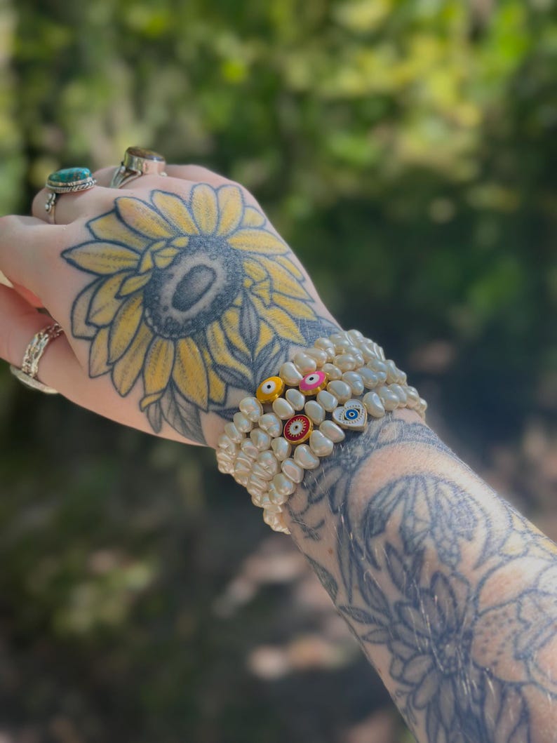 May include: A stack of three pearl bracelets with colorful evil eye beads. The bracelets are made of off-white pearls and feature small, round evil eye charms in various colors. The background is a blurred view of green foliage.