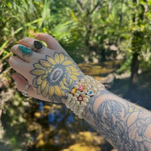 May include: A stack of white pearl bracelets with colorful evil eye and flower accents. The bracelets are worn on a tattooed arm, with rings on the fingers. The background is a blurred view of a natural outdoor setting.