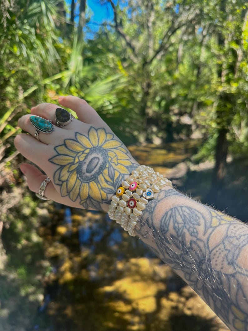 May include: A stack of white pearl bracelets with colorful evil eye and flower accents. The bracelets are worn on a tattooed arm, with rings on the fingers. The background is a blurred view of a natural outdoor setting.