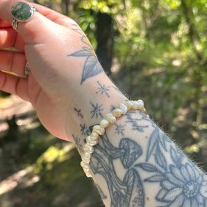 May include: A delicate bracelet made of small, off-white, irregularly shaped pearls. The bracelet is worn on a wrist with tattoos of mushrooms, flowers, and stars. A silver ring with a green stone is also visible.