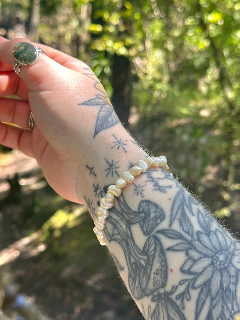 May include: A delicate bracelet made of small, off-white, irregularly shaped pearls. The bracelet is worn on a wrist with tattoos of mushrooms, flowers, and stars. A silver ring with a green stone is also visible.