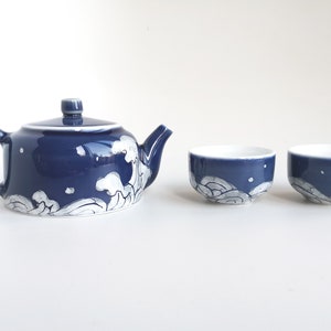 May include: A blue and white ceramic teapot and two matching teacups. The teapot and cups feature a white wave design on a dark blue background.