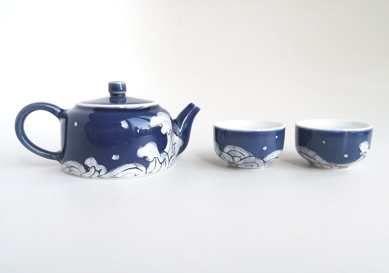 May include: A blue and white ceramic teapot and two matching teacups. The teapot and cups feature a white wave design on a dark blue background.