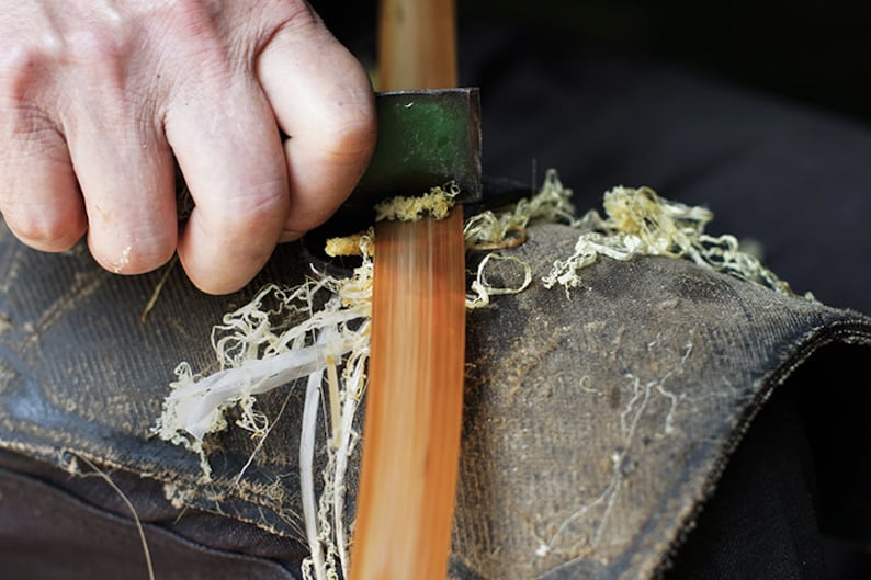 Puede incluir: Una mano usando una peque&ntilde;a herramienta verde para rasurar madera de una pieza larga, delgada y marr&oacute;n de madera. La madera est&aacute; apoyada sobre una superficie de tela oscura con virutas de madera esparcidas alrededor.