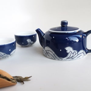 May include: A blue and white ceramic teapot and two matching teacups with a wave design. The teapot has a lid and a handle. The teacups are small and round. There is a wooden scoop with loose leaf tea on a white surface.