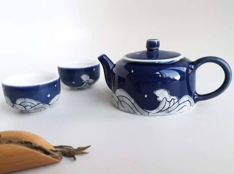 May include: A blue and white ceramic teapot and two matching teacups with a wave design. The teapot has a lid and a handle. The teacups are small and round. There is a wooden scoop with loose leaf tea on a white surface.