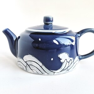 May include: A small blue ceramic teapot with a white wave design. The teapot has a rounded body and a small spout. The handle is on the side of the teapot.