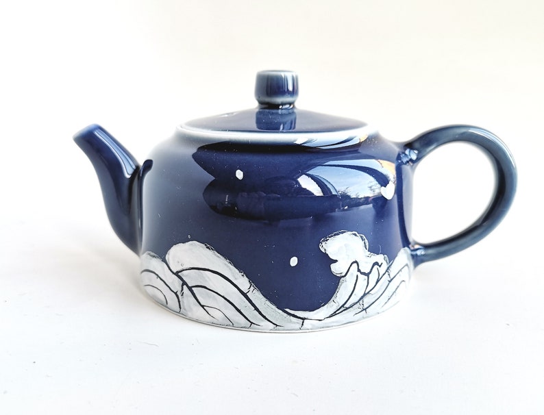 May include: A small blue ceramic teapot with a white wave design. The teapot has a rounded body and a small spout. The handle is on the side of the teapot.