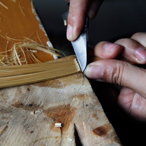 Puede incluir: Se muestran las manos de una persona recortando un haz de finas tiras de bamb&uacute; de color marr&oacute;n claro con un peque&ntilde;o cuchillo. El bamb&uacute; est&aacute; sobre una superficie de madera.