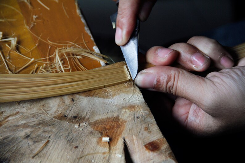 Puede incluir: Se muestran las manos de una persona recortando un haz de finas tiras de bamb&uacute; de color marr&oacute;n claro con un peque&ntilde;o cuchillo. El bamb&uacute; est&aacute; sobre una superficie de madera.