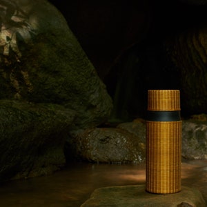Puede incluir: Una botella de agua aislada marr&oacute;n y negra con una textura similar al bamb&uacute; se encuentra sobre una roca en un entorno oscuro y musgoso.