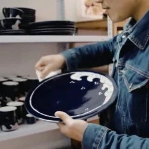 May include: A person holding a blue and white ceramic plate with a wave design. The plate is on a shelf with other blue and white ceramic dishes.