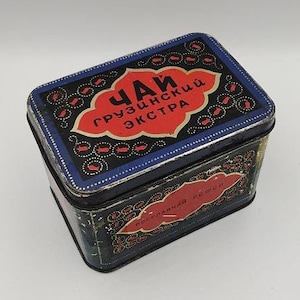 Könnte beinhalten: Eine vintage Metall-Teedose mit schwarzem Hintergrund und blauen und goldenen Akzenten. Der Text auf der Dose lautet "Чай грузинский экстра" in roten Buchstaben.