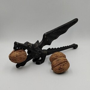 Può includere: Uno schiaccianoci nero a forma di drago, con una noce tra le fauci e due noci vicine. Lo schiaccianoci ha un design dettagliato con ali e una lunga coda. Le noci sono marroni e hanno gusci strutturati.