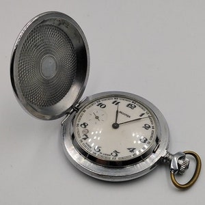 Op de afbeelding: Een zilveren zakhorloge met een witte wijzerplaat en zwarte cijfers. Het horloge heeft een goudkleurige ketting eraan. Het horloge is open en de achterkant is zichtbaar.