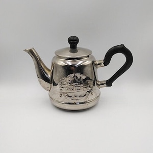 Puede incluir: Una tetera plateada con asa y pomo de tapa negros. La tetera tiene un diseño en relieve en la parte delantera. El pico es curvo y el asa tiene forma redondeada. La tetera es probablemente de metal.