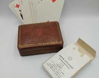 May include: A vintage playing card set with a brown leather case and a partially visible deck of cards. The case has a gold trim and a crest. A partially open card box with Cyrillic text is also present.
