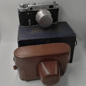 Puede incluir: Una cámara de película ФЭД-2 negra y plateada vintage con una tapa de lente, sobre una caja azul oscuro con texto cirílico. Una funda de cuero marrón está al frente. Un artículo de fotografía clásico.