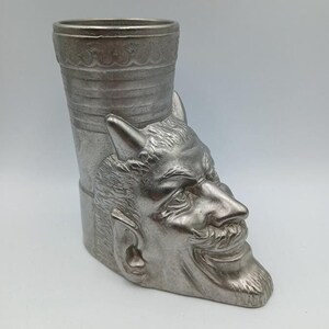 May include: A silver metal cup shaped like a devil's head. The cup has a cylindrical shape and a detailed face with horns, a beard, and a mischievous grin.