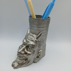 May include: Silver metal desk organizer shaped like a devil's head with horns. The organizer has a cylindrical opening for holding pens and pencils. Three blue and yellow pens are in the organizer.