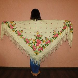 May include: Cream-colored shawl with a floral pattern featuring pink and yellow roses and green leaves. The shawl has a fringed edge and is displayed against a pink wall.
