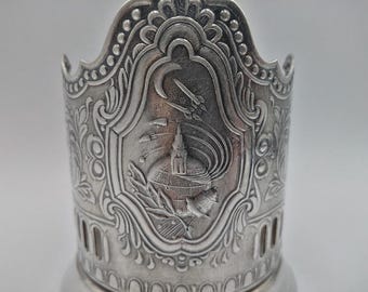 Könnte beinhalten: Silberfarbener Metall-Tassenhalter mit aufwendigen Reliefdetails. Das zentrale Design zeigt einen Turm, Raketen und einen Halbmond. Der Tassenhalter hat einen dekorativen, gewellten oberen Rand und vertikale Aussparungen.