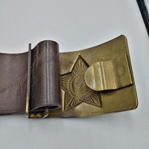 May include: A brown leather belt with a brass buckle featuring a five-pointed star with a circular design in the center.
