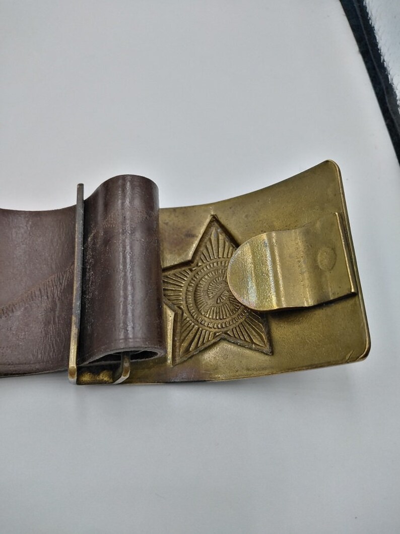 May include: A brown leather belt with a brass buckle featuring a five-pointed star with a circular design in the center.