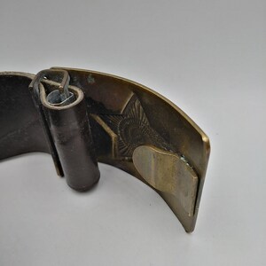 May include: A brown leather belt with a brass buckle featuring a detailed design. The buckle is closed and the belt is folded.