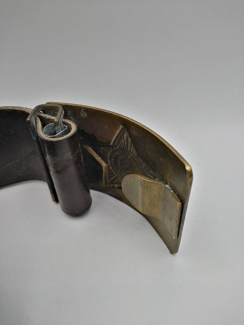 May include: A brown leather belt with a brass buckle featuring a detailed design. The buckle is closed and the belt is folded.