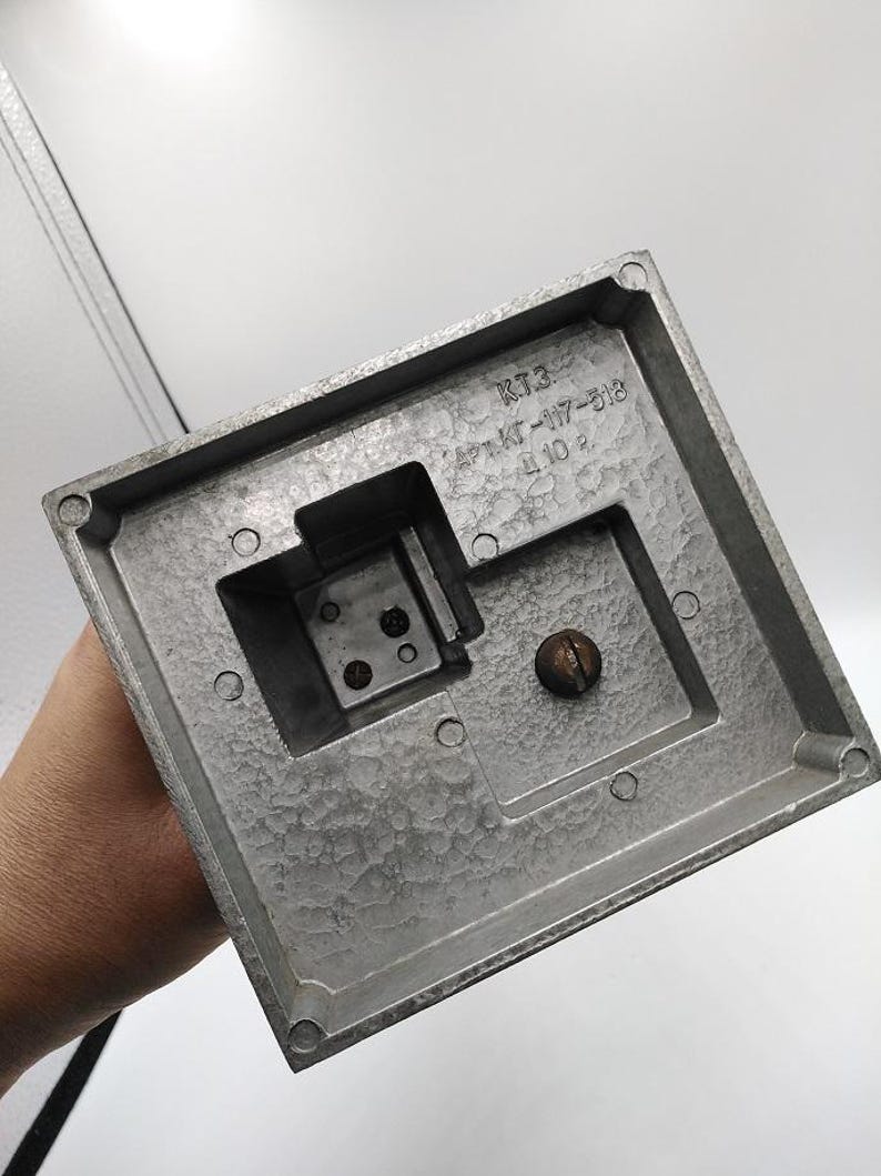 K&ouml;nnte beinhalten: Nahaufnahme eines silberfarbenen, quadratischen Metallobjekts mit strukturierter Oberfl&auml;che. Das Objekt hat eine Aussparung mit Schrauben und einen zentralen, kreisf&ouml;rmigen Knopf. Der Text "KT3" und andere Markierungen sind sichtbar.