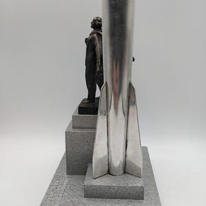 K&ouml;nnte beinhalten: Eine silberfarbene Raketenskulptur mit einer Figur auf einem mehrstufigen Sockel. Die Skulptur hat eine polierte Oberfl&auml;che und ein detailliertes Design. Der Sockel ist grau strukturiert. Die Figur ist dunkelbronzen.