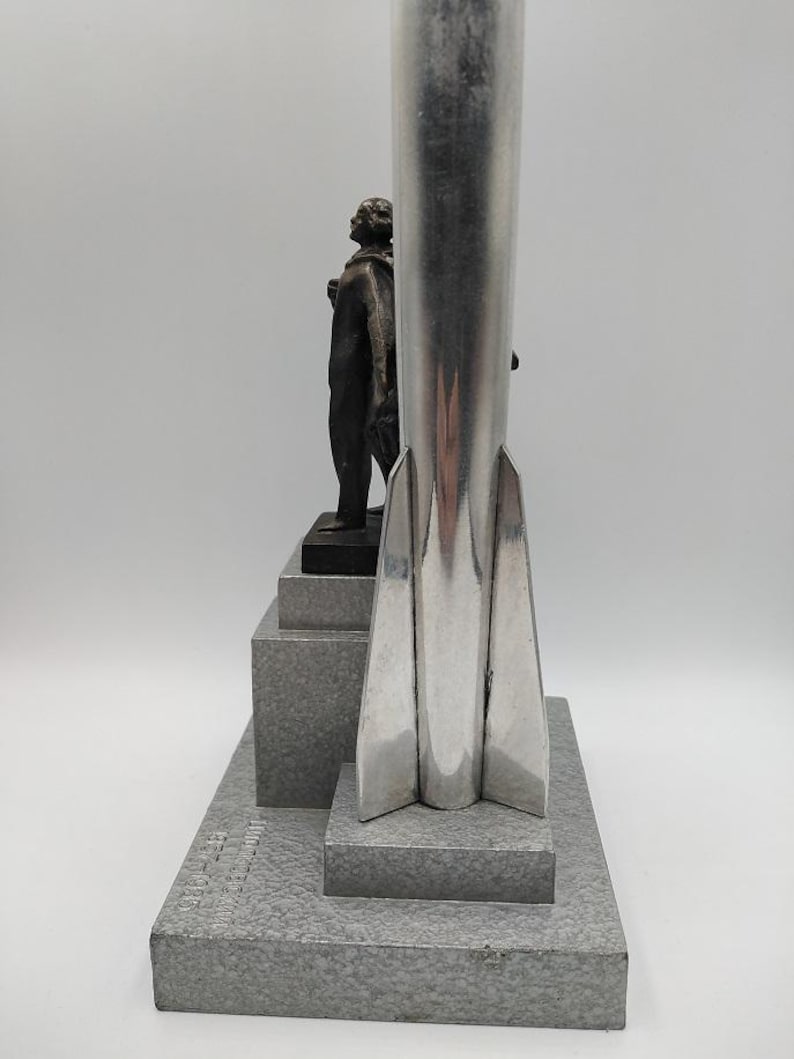 K&ouml;nnte beinhalten: Eine silberfarbene Raketenskulptur mit einer Figur auf einem mehrstufigen Sockel. Die Skulptur hat eine polierte Oberfl&auml;che und ein detailliertes Design. Der Sockel ist grau strukturiert. Die Figur ist dunkelbronzen.